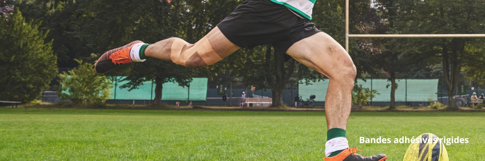 Un joueur de rugby en mouvement frappe le ballon sur un terrain. Elle porte un bandage adhésif élastique sur son genou, illustrant son utilisation pour le soutien des articulations pendant l'activité physique.